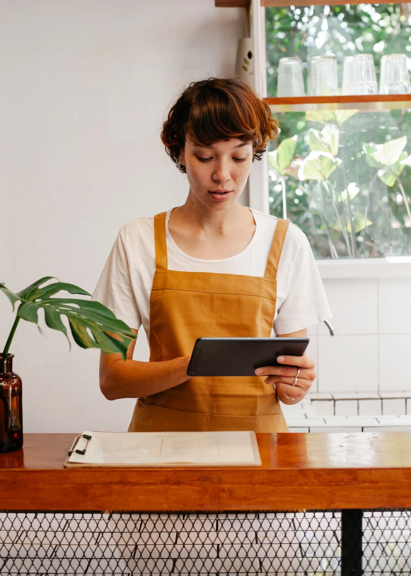 Restaurant employee in an apron using a tablet at a cafe counter, focused on the screen.